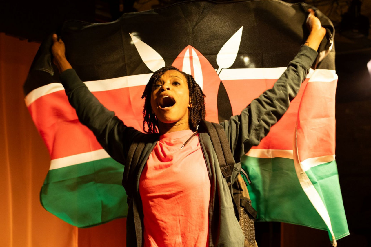 A production photograph from A Fine Idea, showing Grace Saif in character as Kala. She is wearing a black hoody over a red t-shirt,
          and holding aloft a Kenyan flag.