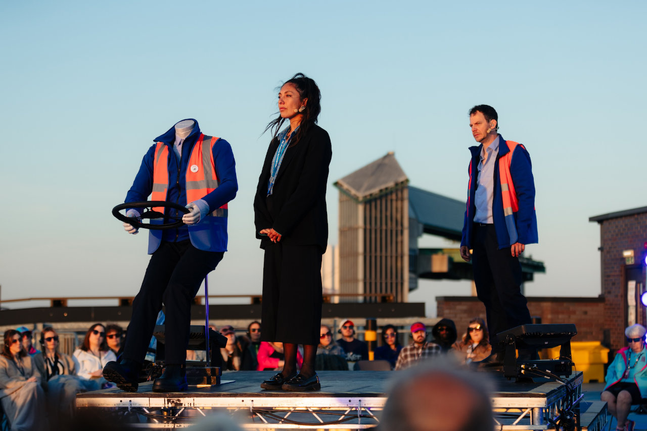 A production photograph from Biting Point, showing the character of Anita, played by Katie Singh, and John, played by Marc Graham.
          Anita is wearing a black suit jacket and cropped trousers, John is wearing a light blue shirt, royal blue fleece and orange hi-vis vest. They are on top of a car park in Hull, the city
          skyline can be seen in the background.