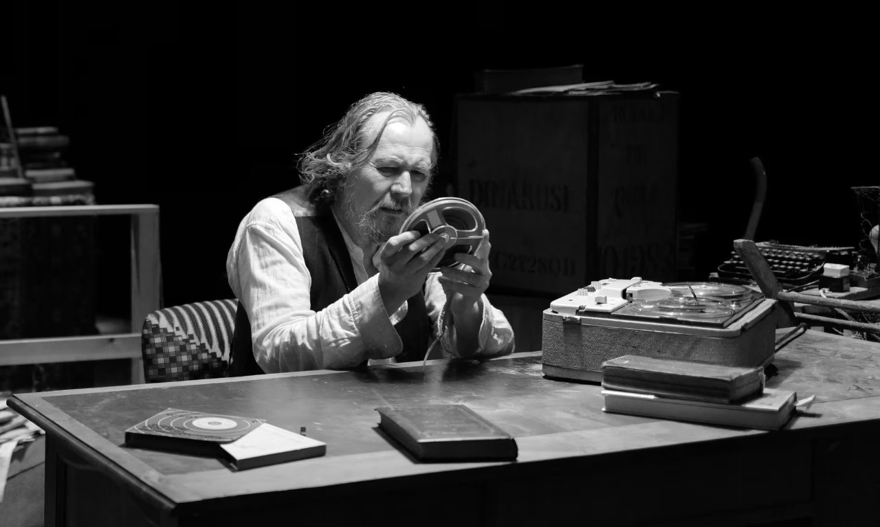 A production photograph from Krapp's Last Tape, showing Gary Oldman in character of Krapp. He is setting at a desk in a cluttered room,
          carefully inspecting a spool of audio tape.