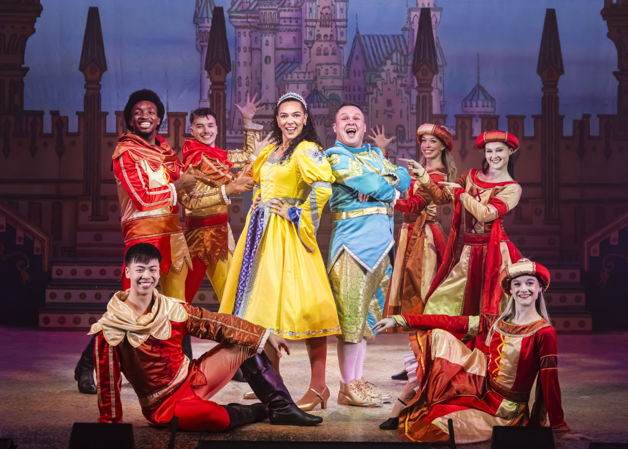 A production photograph from Sleeping Beauty, showing the characters of Aurora, played by Aoife Kenny, and Jangles, played by Tommy Carmichael.
          Aurora is wearing a yellow dress with blue and silver trim, Jangles is wearing a turqoise jacket with gold trim and sparkly gold cropped trousers. They are posing at the end of a musical number, and
          are surrounded by the ensemble, who are wearing red and gold costumes.